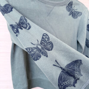 Sleeve detail photo showing alternating moths and butterflies up the sleeve of the olive green sweatshirt, with one moth on the front shoulder.