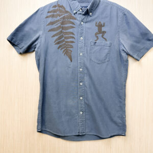 Front of a blue button down printed with a brown fern growing from the right shoulder and a frog print jumping out of the left pocket. Some slight lightening under armpits.