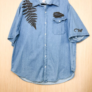 Front of blue short-sleeve button down shirt with brown fern print growing down from right shoulder, oyster mushroom printed growing out of left pocket, and long-toed salamander printed on right sleeve.