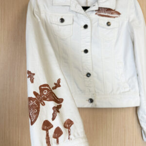 Hand holding sleeve of white denim jacket in foreground showing print of moth, three bats, and three LBMs in brown ink.