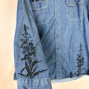 Hand holding sleeve of denim button down shirt with morels printed on right breast pocket, fireweed on right sleeve, and fireweed and morels along lower left hem.