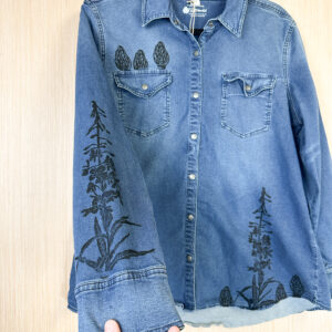 Hand holding right sleeve of denim button down shirt with morels printed on right breast pocket, fireweed on right sleeve, and fireweed and morels along lower left hem.