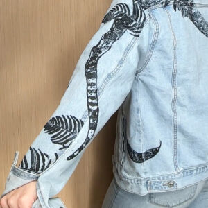 Back of woman wearing denim jacket with left arm printed with bull snake and ferns in black ink, head of snake poking out of left pocket.