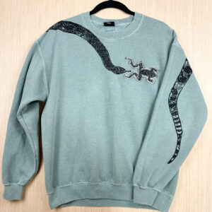 Front of light blue crewneck sweatshirt with black snake coming down from right shoulder, denim frog patch on left breast right in front of snake's mouth, tail print shown on back side of folded over left sleeve.