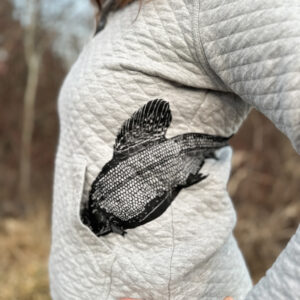 Front left of woman wearing light gray quilted sweatshirt, arctic grayling print nose first in front pocket, wrapping up and around torso.
