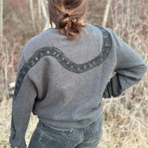 Back of woman wearing grey sweatshirt, showing black snake print draping down from right shoulder, around the back in a u shape, and tail down left sleeve.