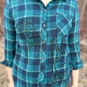 Front of woman wearing a plaid flannel button down with steelhead head printed coming up from right hem, ending at breast pocket, tail starting at right breast and wrapping around right side.