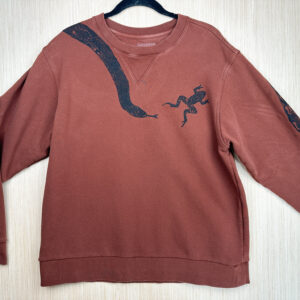 Front of burnt orange sweatshirt with black snake print coming down from right shoulder, frog printed jumping away from snake tongue on left breast, edge of tail print visible on top of left sleeve.