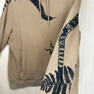 Sleeve of light brown hoodie held in foreground, with fern and snake tail printed in black ink going up back of sleeve, snake head printed across chest visible in background, with fern growing down right sleeve and frog printed jumping away from snake head.