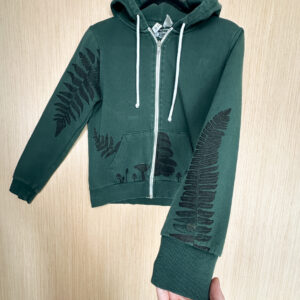 Hand holding left sleeve of green sweatshirt in foreground, printed with black sword fern, pockets printed with mushrooms, and a bracken fern printed growing out of right pocket and down right sleeve from shoulder seam.