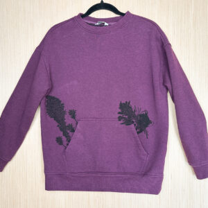 Front of berry color crewneck sweatshirt with front pocket printed in black ink with a life-size pika popping out of left side of pocket with leaves in its mouth, beargrass and indian paintbrush prints growing out of other side.