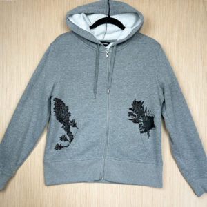Front of grey zip hoodie with life size pika printed in black ink popping out of left pocket, beargrass and three indian paintbrush flowers printed growing out of right pocket.