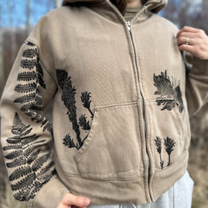 Front of cropped tan zip hoodie, with black ferns on right sleeve, flowers coming out of right pocket, and pika coming out of left pocket.