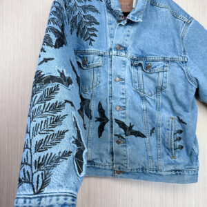 Hand holding sleeve of blue denim jacket in foreground with bat prints up right sleeve and across front of body, with cedar branches down right sleeve, down from right shoulder, and mushrooms coming out of front left pocket.