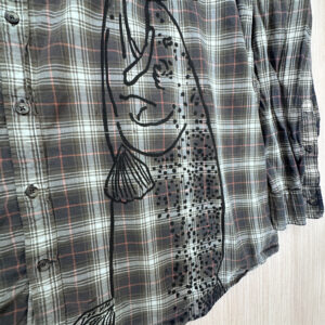 View of black steelhead head print coming up from bottom left hem of brown plaid button up.