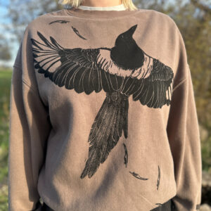 Blond woman wearing tan/brown crewneck sweatshirt with life-size black billed magpie with wings spread printed across the chest, tiny primary feathers printed in black across the front from right shoulder to bottom left hem.