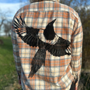 Back of blond woman wearing peach-white-and-blue plaid flannel showing life-size black-billed magpie with wings spread across the back of the shirt.