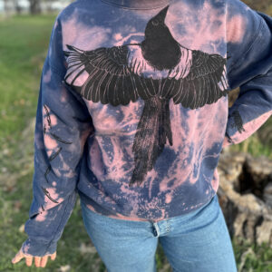 Woman wearing blue sweatshirt with pink bleach design across chest and sleeves, printed with black-billed magpie across chest, tiny wing feathers across right sleeve.