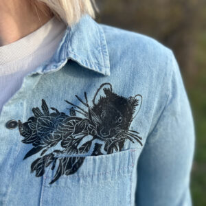 Closeup on left breast pocket of blond woman wearing denim button down with life-size pika printed in left breast pocket.