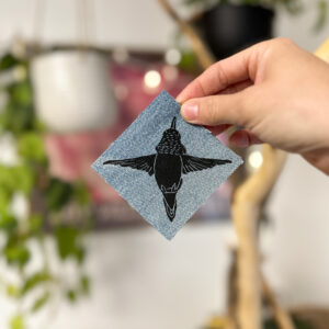 Hand holding a denim sticker that's a square shape in a diamond orientation with a black rufous hummingbird on it