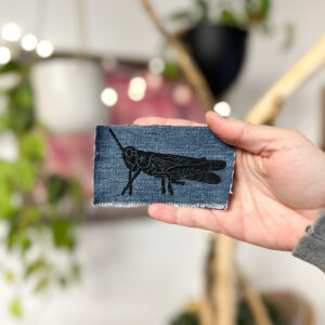 Hand holding a denim sticker with a black print of a grasshopper on it.
