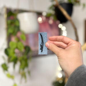 Denim fabric sticker with life size magpie primary feather printed on it in black ink.