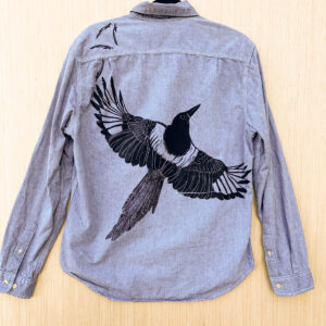 Back of light grey button down with black-billed magpie printed with wings spread across the back at an angle, flying to the right, a flurry of tiny primary feathers printed coming down from left shoulder.