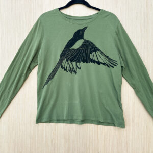 Front of green long sleeve t-shirt printed with flapping black-billed magpie printed in black ink at an angle across chest.