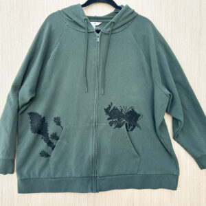 Front of green zip hoodie with raglan sleeves with life size pika printed in black ink popping out of left pocket with crease in leaf print held in its mouth, beargrass and three indian paintbrush flowers printed growing out of right pocket.