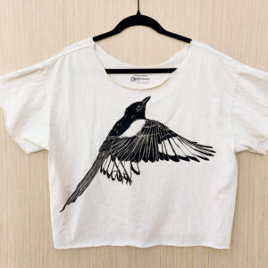 Front of off-white cropped t-shirt with raw edge at hem and sleeves, with ripped collar and life-sized flapping black-billed magpie printed in black ink.