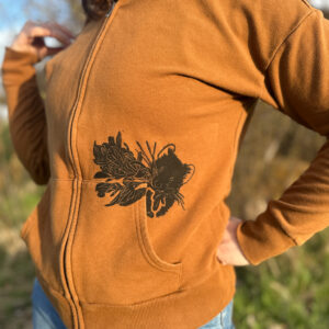 Front of woman wearing golden-brown zip hoodie, showing pika printed coming out of left pocket holding leaves in its mouth.