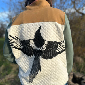 Back of brunette woman wearing quarter-button sweatshirt with quilted off-white body, brown shoulder and green sleeves with a life-size black-billed magpie with its wings spread printed across the back in the off-white block.