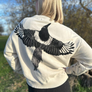 Back of blond woman wearing off-white sweatshirt with a life-size black-billed magpie with its wings spread printed across the back.
