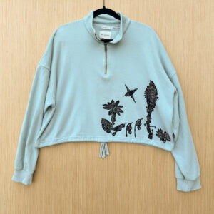 Front of blue cropped quarter zip sweatshirt printed with heartleaf arnica, glacier lilies, beargrass, and bitterroot growing up from lower left hem in black ink with rufous hummingbird pollinating heartleaf arnica.