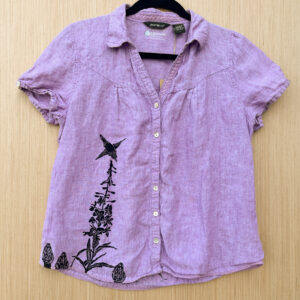 Front of pink short sleeve blouse, printed in black ink along lower right hem with fireweed and three morels, with a rufous hummingbird pollinating the fireweed.