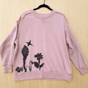 Front of pink sweatshirt with heartleaf arnica, yellow bell, beargrass, and bitterroot printed growing up from lower right hem, with rufous hummingbird pollinating the beargrass.