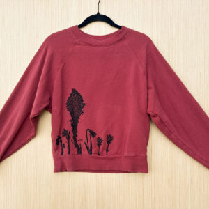 Front of berry colored sweatshirt with ghost pipe, beargrass, yellow bell, and Indian paintbrush printed along lower right hem.
