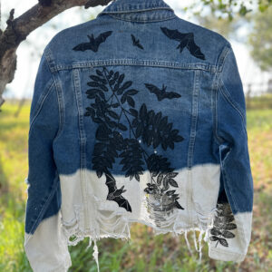 Back of blue denim jacket bleached white and frayed along hem, printed with bats and mountain ash branches.